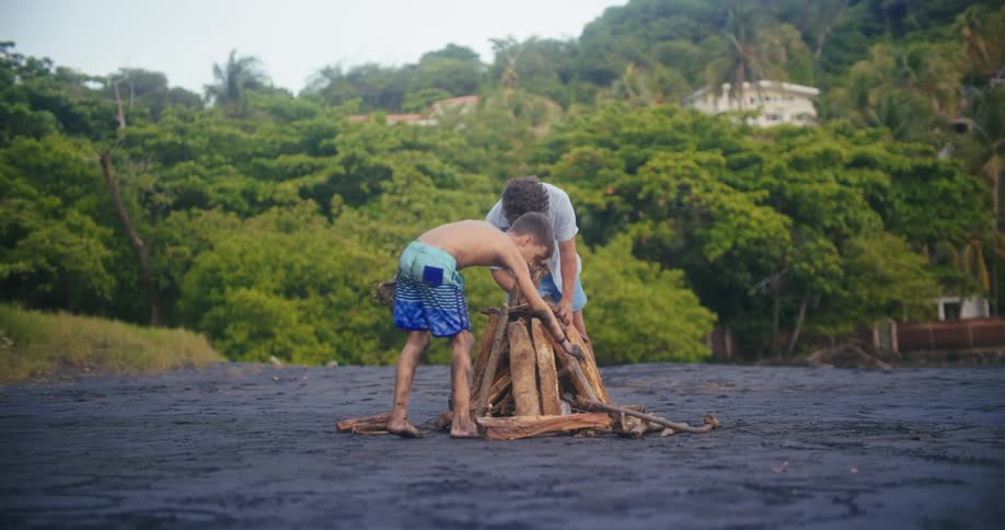 Two young boys one with a pile of logs gather around a fire pit on a beach with dark sand preparing to build a bonfire they work together to arrange the wood with a lush green hillside in the background