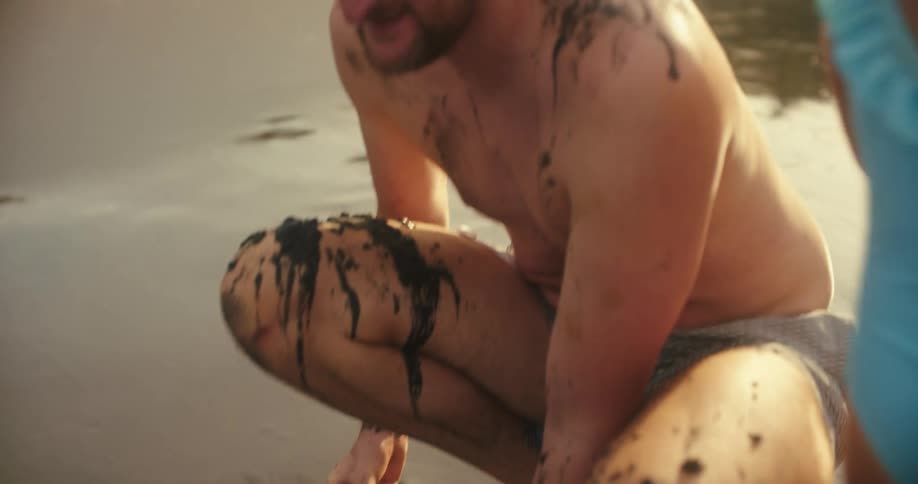 A little girl in a blue and white swimsuit is playing with dark sand on a beach she looks up and smiles at a man kneeling in front of her whose face and hair are covered in the same dark mud