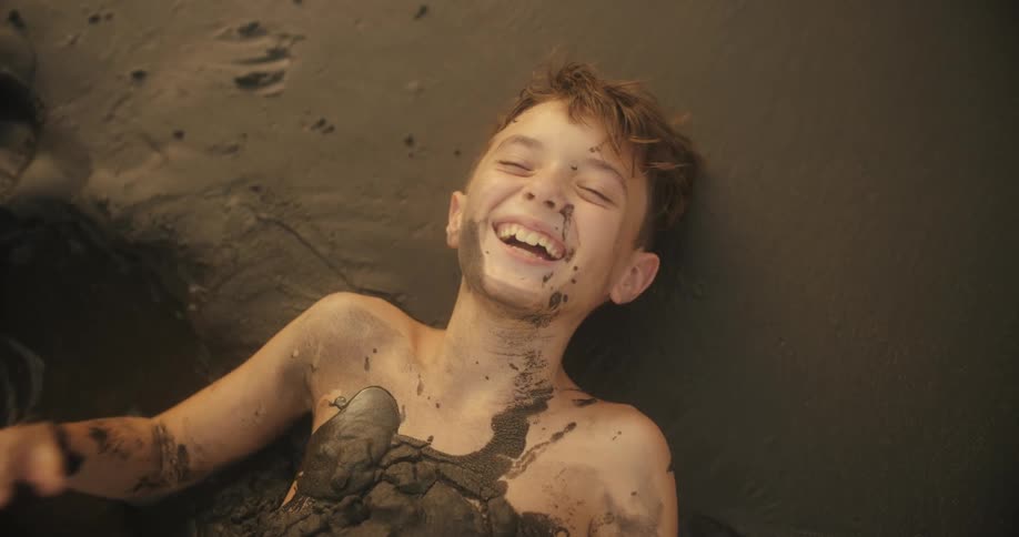 A child with mud on their body lies down and laughs while looking up