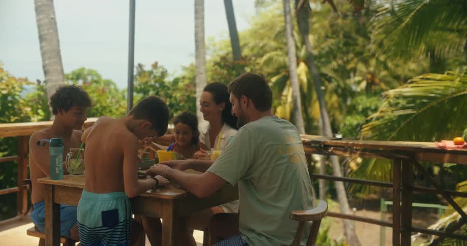 A family enjoys vacation time at a wooden table under palm trees