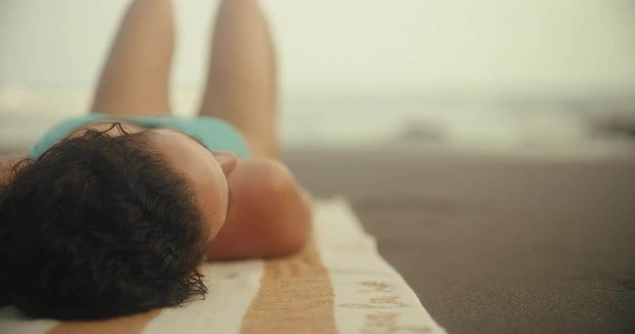 A woman relaxes on a towel at the beach facing the ocean