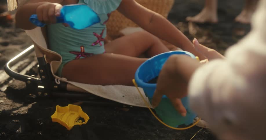 A small girl in a light blue swimsuit sits on a canvas beach chair and uses a blue shovel to scoop sand from the ground into a matching bucket