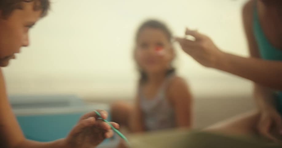 A young girl is sitting on a beach and being fed a strawberry by a woman with a fork with another boy sitting nearby with a cooler