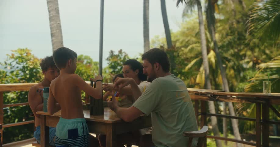 A family is gathered around a wooden table on a tropical sunlit balcony playing a block stacking game together