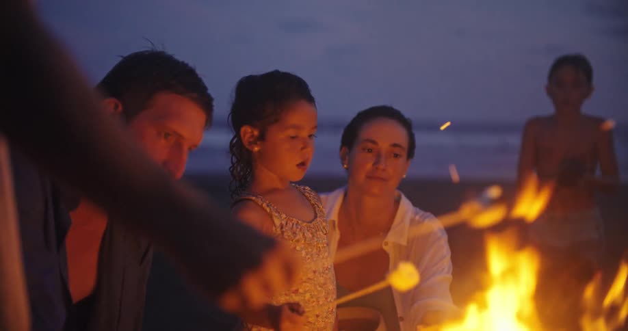 The video shows a family of three a man a woman and a young girl gathered around a fire on a beach at night they are roasting marshmallows over the fire