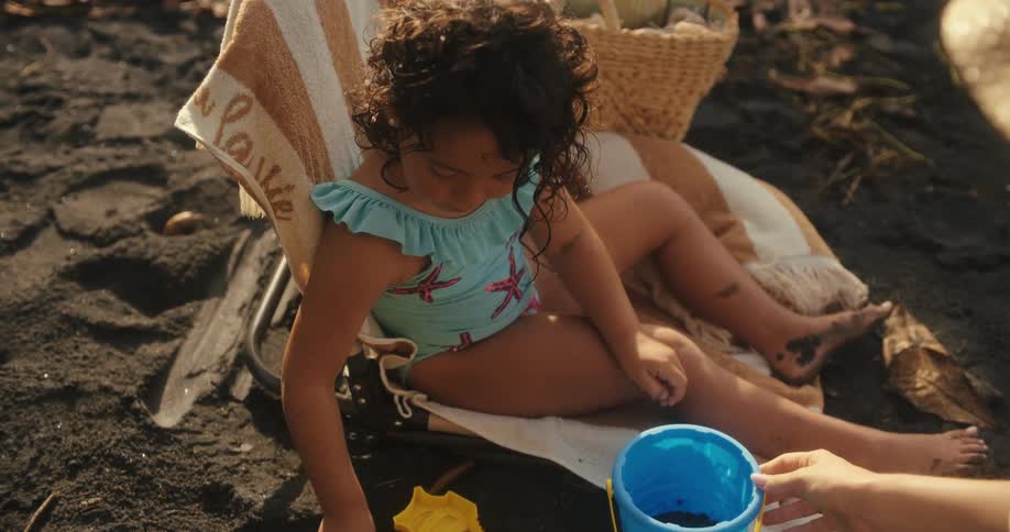 A child sits on a beach and scoops sand with a shovel to place it into a bucket held by another person