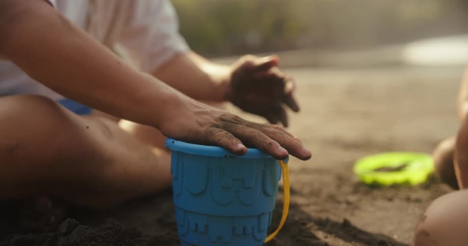 Hands press sand into a blue bucket on the beach