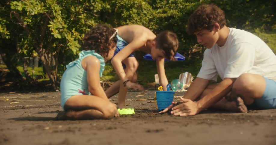 A family of three children plays together on a dark sand beach with lush green foliage in the background they are focused on playing with sand toys building and digging in the sand together