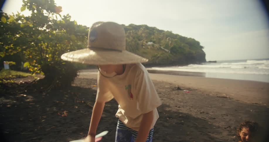 A child with a large straw hat and a backpack walks on a dark sand beach holding a boomerang a younger child sits nearby on a towel playing with a bucket and toys