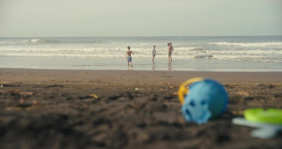 The video shows two children and an adult playing in the water at a sandy beach one of the children is running towards the other two who are standing in the shallow water with waves in the background