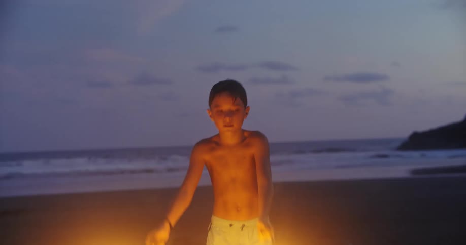 Boy playing with sparklers on sandy beach near shoreline at dusk