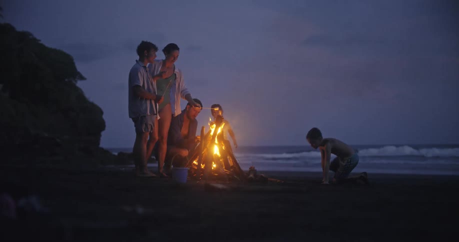 A family gathers around a bonfire on the beach at dusk