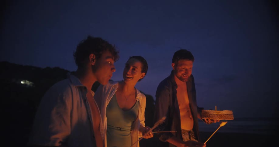 A shot focuses on a small group of people standing on a beach at night with the sky transitioning from sunset to dark they are gathered around a small bonfire roasting marshmallows and enjoying the cool air