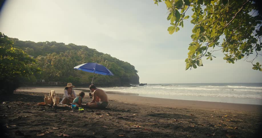 A mother and father sit on a beach with their young child under an umbrella