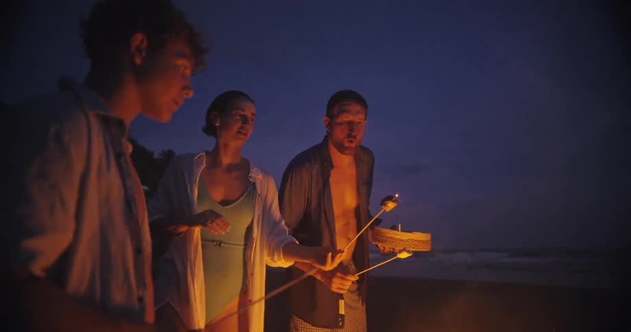 Three people stand around a fire on a beach at night and hold sticks with marshmallows over the flames
