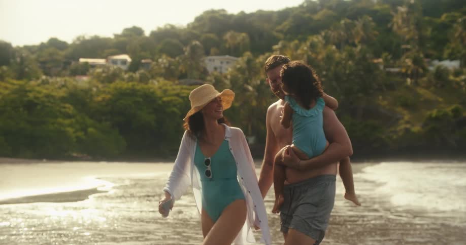 On a bay beach a woman calls her family out from the water and as they come out a man carrying a little girl splashes the woman with water as they all laugh