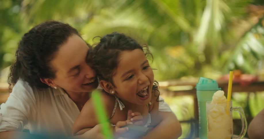 The video shows a woman and a young child sitting at a table with green foliage in the background they are looking down and appear to be engaged in an activity and the woman smiles as she looks at the child