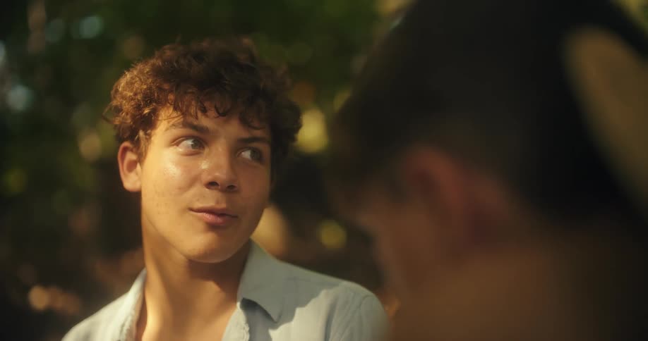 A young man with curly brown hair and bright green eyes looks at an unseen person offcamera he appears engaged and thoughtful while listening to them speak