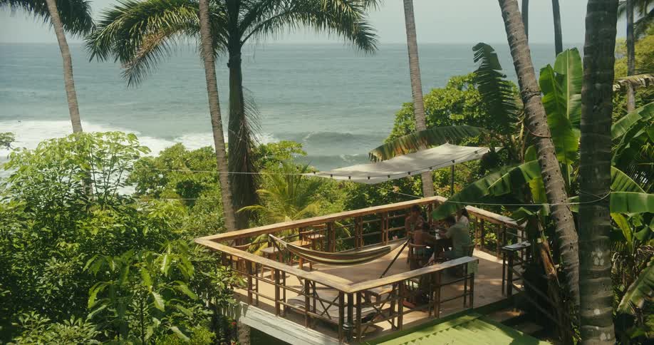 A family talking while seated at a wooden table on a large deck with a hammock and a white canopy all surrounded by dense tropical foliage with an ocean and a white sandy beach in the background