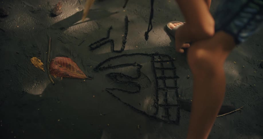 A young boy uses a stick to draw some shapes in the sand