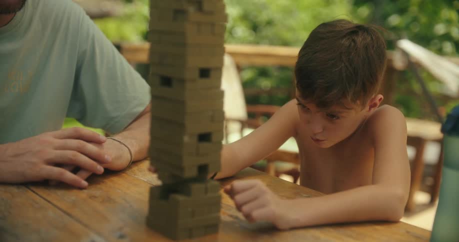 A child with light brown hair sits at a wooden table across from a man leaning forward with a focused expression to remove a block from a tall stack of brown blocks