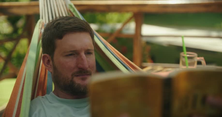 A man with a beard relaxes in a colorful hammock engrossed in reading a book on a sunny day he turns a page seemingly lost in the story and at peace with his surroundings