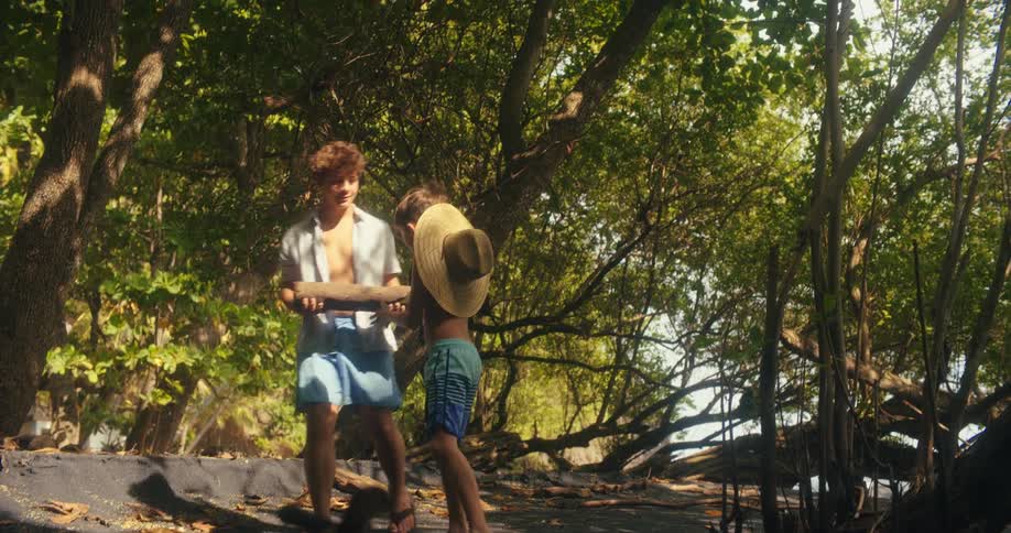Two boys walk on sand in a forest and one of the boys picks up a piece of wood