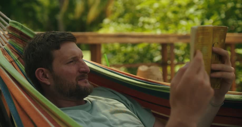 A man with a beard lounges comfortably in a striped hammock his focus fixed on a paperback book he turns the pages slowly his expression one of calm concentration