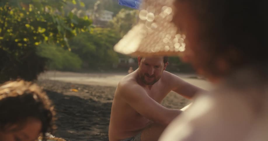 A family is on a sunny beach where a man is seen putting a striped towel on the ground and a woman with a wide brim straw hat is looking at a young girl in a light blue swimsuit