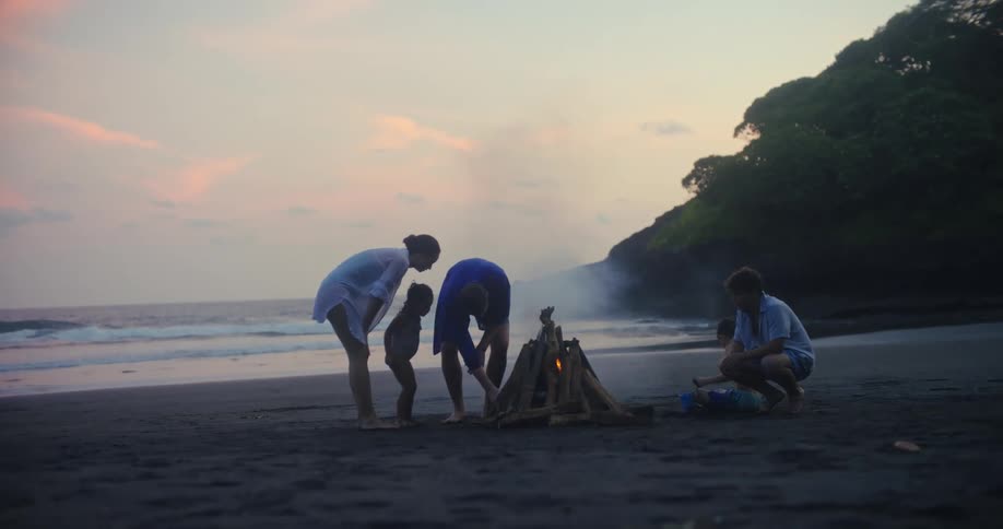 Father and mother with three kids of varying ages lighting a bonfire at the beach next to the ocean outdoors during dusk or dawn