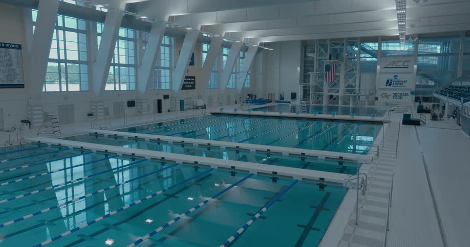 A shot captures the view of an olympicsized indoor pool focusing on the calm lightblue water the pool is empty and illuminated by sunlight streaming through large windows