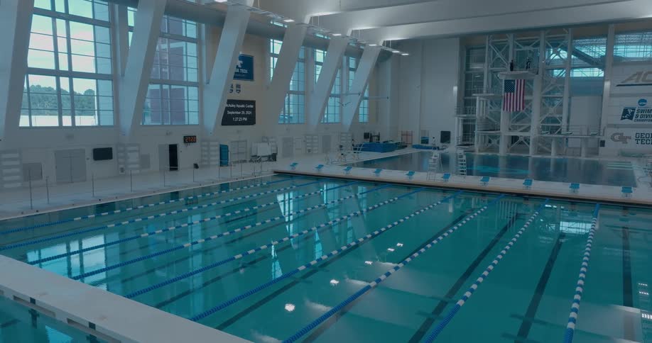 A shot captures the expansive view of a large indoor swimming pool empty and serene the camera slowly pans across the quiet space highlighting the blue lane lines and diving boards against the vast sunlit windows