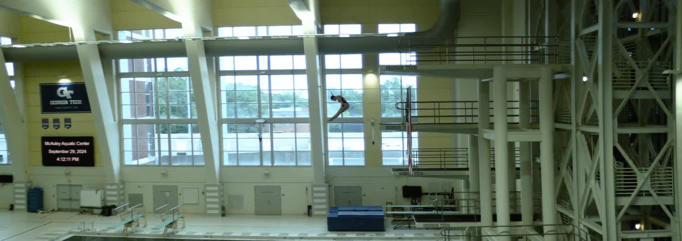 A man in a dark swimsuit with a white trim performs a dive in a large indoor pool facility with high ceilings and large windows beginning his fall from the highest platform of the tall diving structure