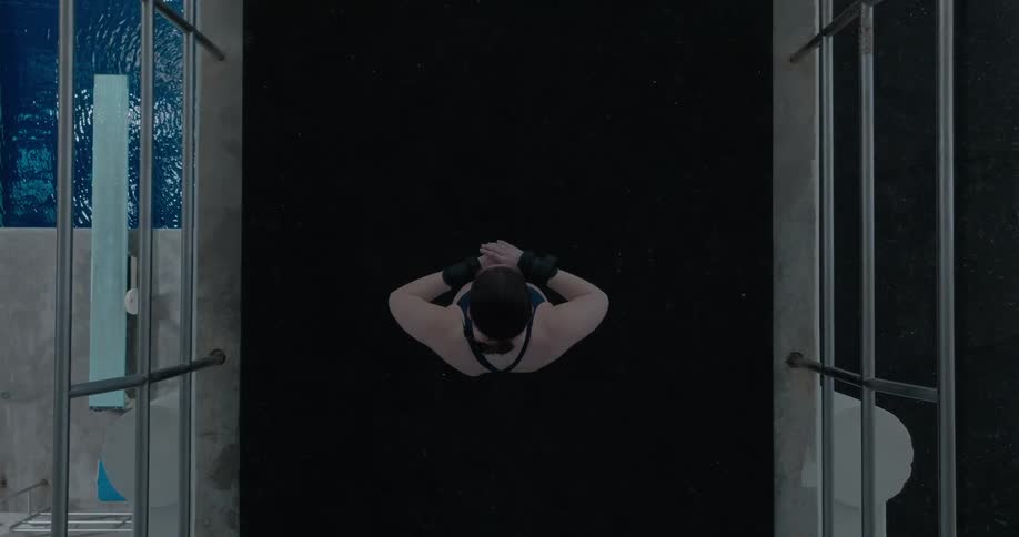 A woman in athletic wear raises her arms while standing on a diving platform above a pool