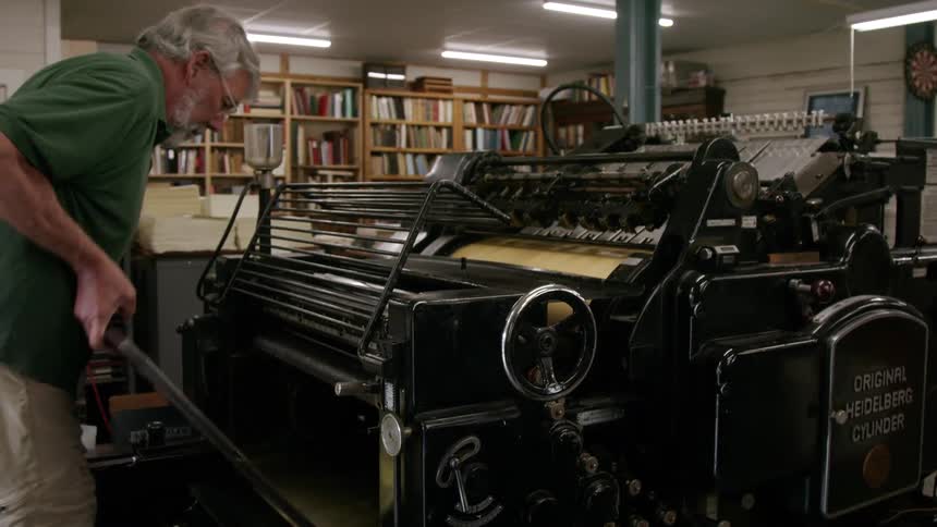 Older man uses a printing press containing an original heidelberg cylinder