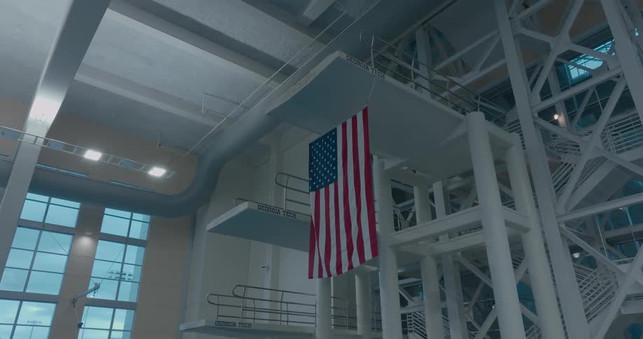 A wide view shows a large american flag hanging down from a tall diving platform structure in a big empty indoor swimming facility