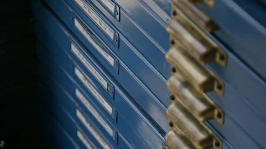 A close view of a set of blue drawers in a storage cabinet with brass handles and white labels is shown as a hand pulls one of the drawers open