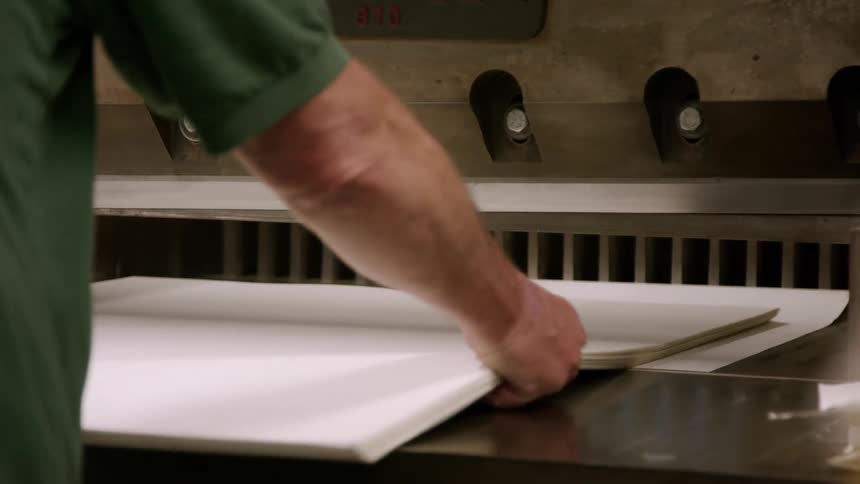 A man wearing a dark green shirt places a stack of large white papers into a wide paper cutting machine