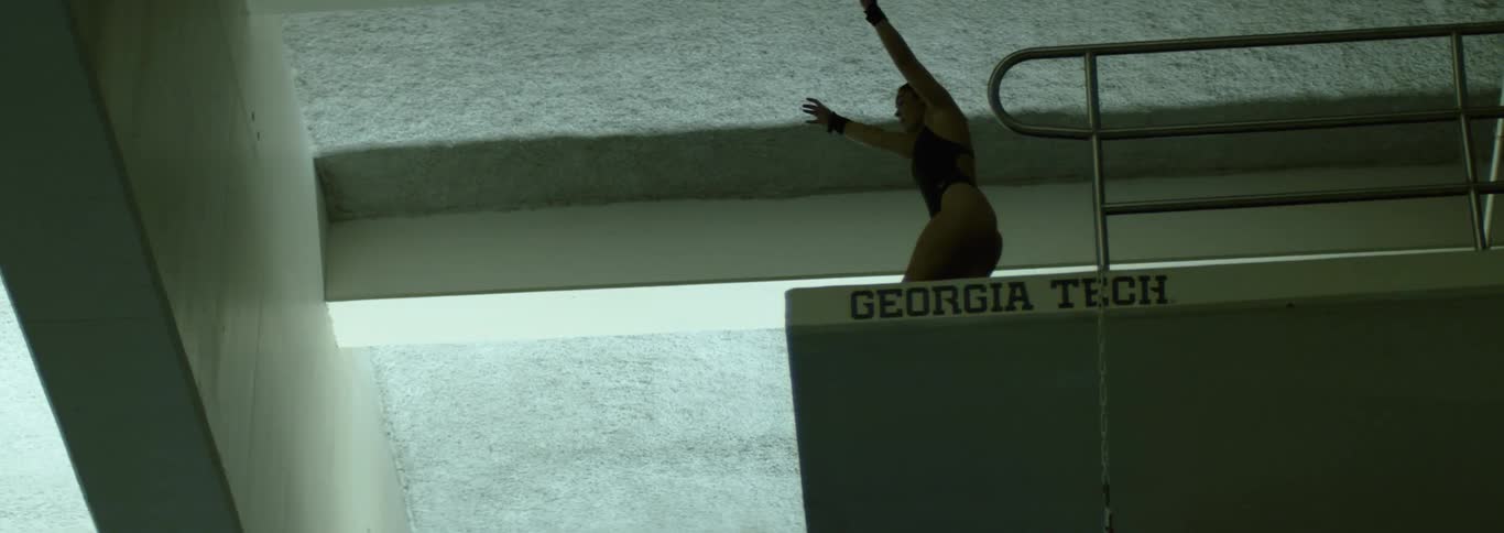 A shot focuses on a female diver sitting on a diving board preparing to perform a dive she holds her arms over her head and rocks slightly focusing on the water below