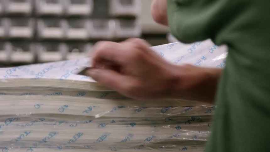 The video shows a closeup shot of a persons hands as they open a large stack of papers wrapped in plastic using a box cutter they slice through the top layer of plastic to unseal the bundle