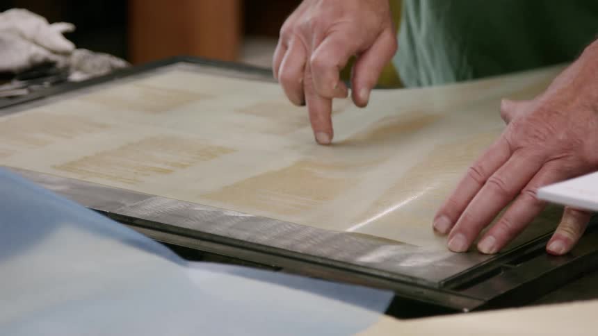 A printer is seen using a handheld tool to apply pressure to a surface covered with a blue textured material the printer then lifts the material revealing a beige sheet underneath with faint pressedin markings