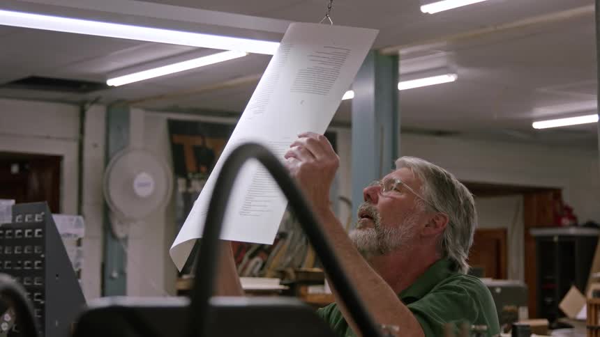 An older man with gray hair and a beard holds up a large printed sheet under bright lights carefully examining the text