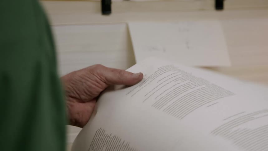 The video shows an older man with gray hair and glasses working on a machine he is focused on the task at hand which involves a series of small metallic levers and a sheet of white paper