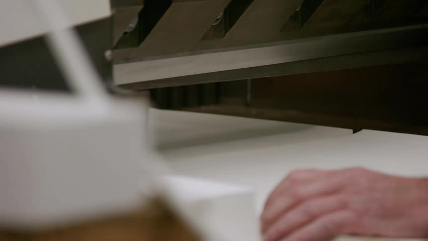 The video shows a closeup shot of a paper cutter in a manufacturing or printing facility white paper is fed into the machine which then cuts a section off