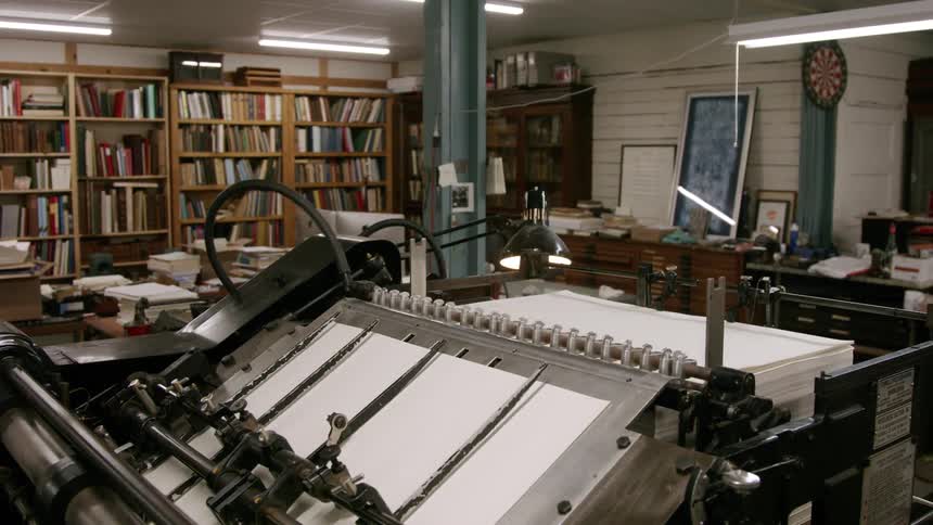 In the foreground of the video a large silvercolored printing press is actively processing sheets of paper in the background theres a busy workshop space filled with bookshelves various work surfaces and what appears to be a variety of tools and e