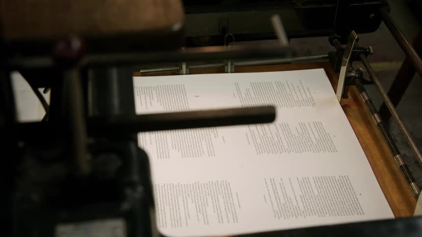 A persons hand quickly removes a printed sheet of paper from the bed of a large printing press