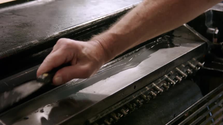 A man is using a tool to apply a thick dark substance onto a printing press roller he uses a small spatula to spread the ink evenly across the roller