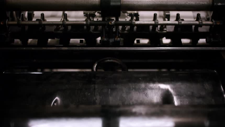 A sheet of white paper with black text moves quickly through the interior of a large dark printing press machine which is filled with many metal parts