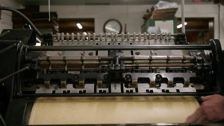 A person operates a vintage printing press carefully placing a large sheet of paper onto a roller before the complex machinery begins its cycle