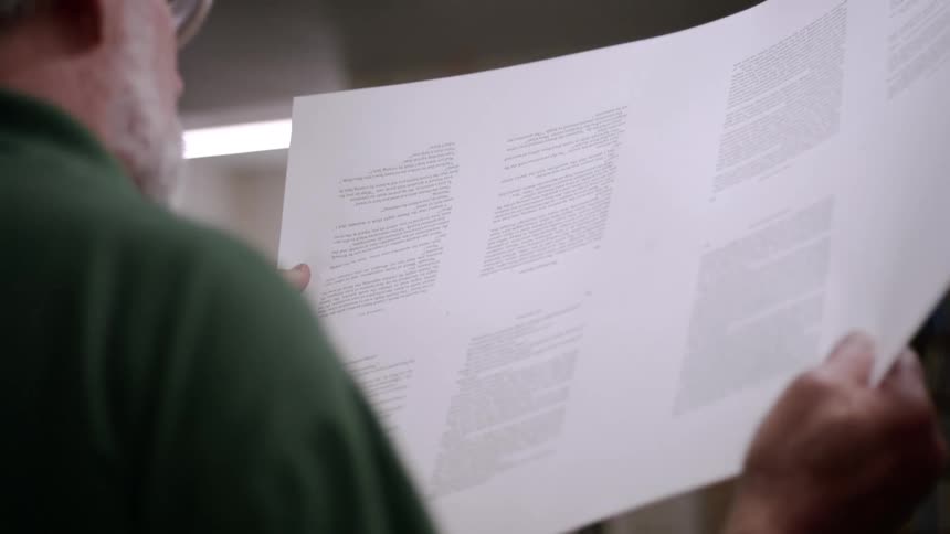 A printer holds up a large sheet of paper examining it closely the paper is filled with dense text and the printers face is obscured by his glasses and the sheet